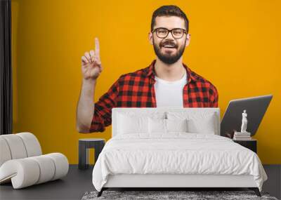 Young bearded man holds laptop and finger point up isolated against yellow background. Wall mural