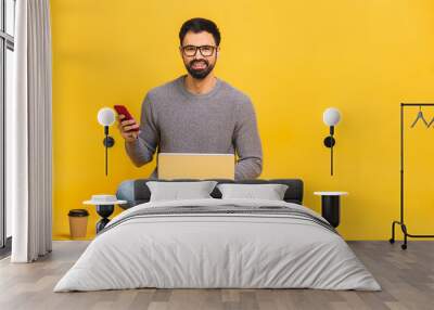 Young bearded man sitting on the floor with laptop and talking at phone. Isolated over yellow background. Wall mural