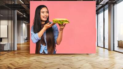 Young beautiful asian japanese chinese woman eating sandwich or big burger with satisfaction. Girl enjoys tasty hamburger takeaway, diet concept, standing isolated over pink background. Wall mural