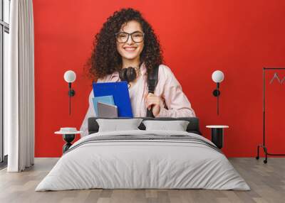 Young curly student woman wearing backpack glasses holding books and tablet over isolated red background. Wall mural