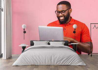 Young smiling african american man standing and using laptop computer isolated over pink background. Wall mural