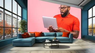 Young surprised shocked amazed african man standing and using laptop computer isolated over pink background. Wall mural