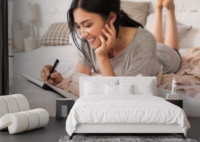 Beautiful young asian woman laying on bed and writing a diary. Smiling brunette lady with notebook and pen in her hands. Modern bright home interior on background. Wall mural