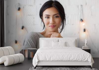 Beautiful young asian woman with brunete hair looks at camera. Cute girl posing in front of white brick wall. Smiling lady in casual dress, white wall with light bulbs on background. Wall mural