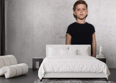 Cute little boy in black T-shirt posing in front of grunge concrete wall. Portrait of fashionable male child, copy space. Boy looks at camera, gray wall on background. Wall mural