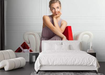 Portrait of attractive cheerful girl with playful look. Blonde hair beautiful woman sits in white armchair with present in her hands. Young woman portrait hold gift in christmas color style. Wall mural