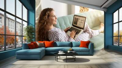 Portrait of beautiful smiling girl doing shopping on internet at home. Cheerful lady laying on sofa in pretty pyjamas. Morning and technology concept. Blurred background Wall mural