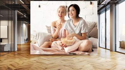 Portrait of funny and happy young women watching comedy in bed and laughing. Cheerful friends eating tasty popcorn and looking movie with gladness. Cozy and friendly atmosphere Wall mural