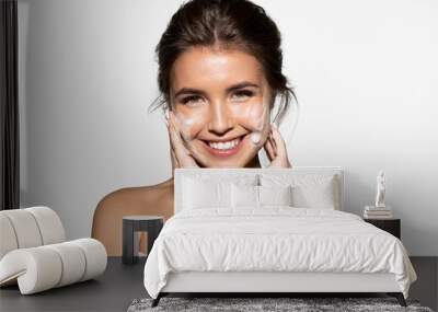 Portrait of joyful young woman applying white foam for washing. Cheerful brunette with perfect clean skin looking at camera with gladness. Skincare spa relax concept. Isolated on grey background Wall mural