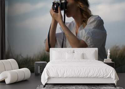 attractive caucasian woman with film camera in white sundress sitting on the meadow capturing the landscape against the blue sky Wall mural