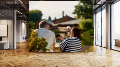 Back view sitting man and woman outdoors. mature woman shows something funny on her phone to her husband. Wall mural