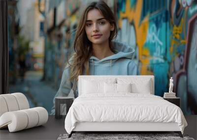 Young woman in a casual hooded sweatshirt, holding a cotton tote bag mockup, standing against a graffiti wall in the city Wall mural