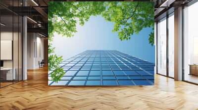 A low-angle view of a modern glass skyscraper with green tree branches overhead against a bright blue sky. Wall mural