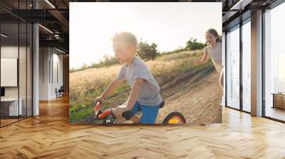 Boy and girl in the field. The boy is riding a bicycle, and the girl is running alongside. A cheerful, happy childhood in the village. Wall mural