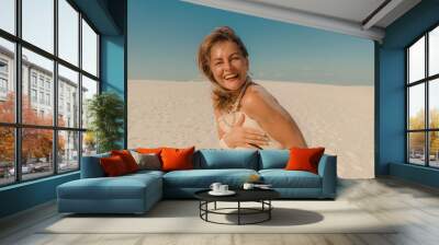 Laughing mature woman with no bikini holding summer hat on sandy beach Wall mural