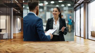 Two business professionals, a man and a woman, engage in a lively discussion while standing in a busy factory hall, demonstrating collaboration and industrial success. Wall mural