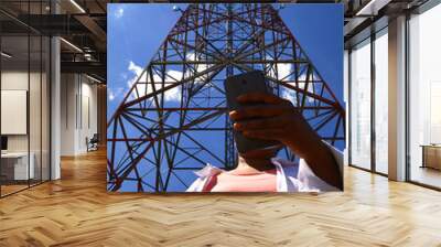 Hands of a woman holding a smartphone. Looking for wi fi connection. Online social activity and connection concept.  Very sunny day. Antenna, tower mobile communication and blue sky behind. Wall mural