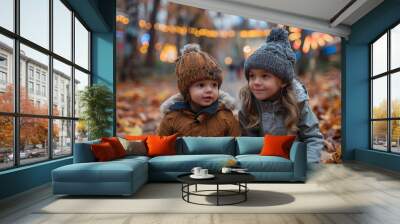 Sitting on a bed of fallen leaves, two toddlers share a moment of enjoyment with an amusement park in the background Wall mural