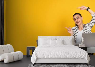 Woman pointing copy space, young happy cheerful girl wear casual shirt sit at office desk woman pointing copy space. Using laptop fingers showing aside mock up area. Isolated yellow background.  Wall mural