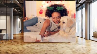 Cute young woman wearing a colorful green set of Christmas reindeer antlers taking a selfie with her dog as they relax in the living room in front of the fire. Wall mural