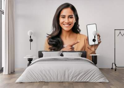 A young modern happy Indian Asian woman or female is showing and pointing a finger at a mobile phone or smartphone blank screen and smiling looking at the camera isolated on a white background. Wall mural