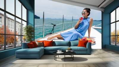 A beautiful asian lady in a blue dress on a yacht drinks champagne and eats fruit, on the background of sea Wall mural