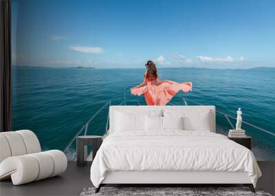 Outdoor shot of adorable young woman in a pink dress on edge of yacht, looking to beautiful nature landscape during trip. Happy woman enjoying summer travel. Back view Wall mural