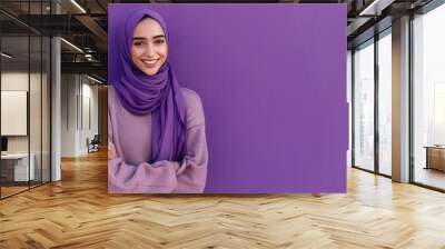 A portrait of a young smiling Muslim woman wearing a purple hijab against a purple background studio with copy space for text Wall mural