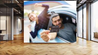 Happy cheerful young couple traveling by the car, male car driver showing a credit card to camera. Concept of travel and transportation insurance. Man paying a gas or EV charging by credit card. Wall mural