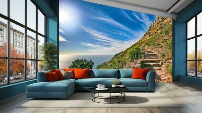 A mountain trail with blue sky and lensflare over ocean. Landscape of mysterious dirt road for hiking on adventure walks along a beautiful scenic trail with lush shrubs in Cape Town, South Africa Wall mural