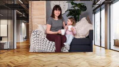 young woman and her cute little daughter sitting and drinking tea in living room Wall mural