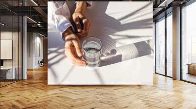 Woman's hands and a glass of clean water on the white table in natural sunlight with plant shadows. Wall mural