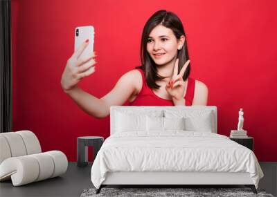 Cool classy woman in red t-shirt partying and taking selfie with victory sign on her cell phone isolated over red background Wall mural