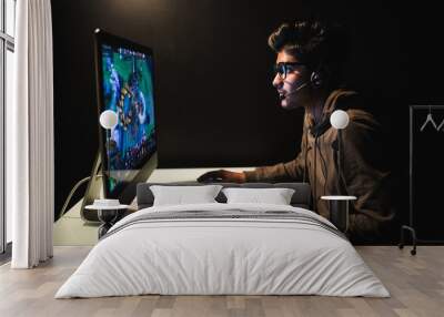 Gamer or streamer in earphones with microphone sits at home in dark room and plays with friends on networks in video games. A young man sits in front of a monitor. Wall mural