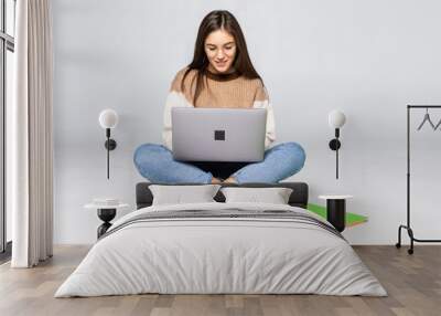 Happy young woman sitting on the floor with books and using laptop on white background Wall mural