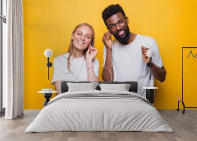 Portrait of joyful couple african man and caucsian woman singing while using smartphone and earphones together over yellow background Wall mural