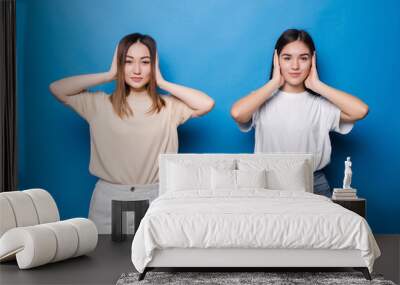 Portrait of two funny mixed race women covering ears isolated over blue background Wall mural