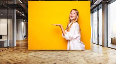 Young happy woman holding her hand palm up, ready to hold your product on yellow background Wall mural