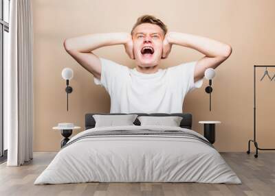 Young man covering his ears with hands opens his mouth screaming isolated on a beige background. Wall mural