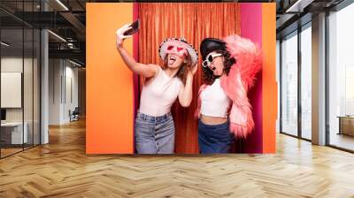 Attractive couple of casual caucasian women posing with carnival dress having fun standing with yellow - orange background hugging and making face..Two ladies making a selfie. Wall mural