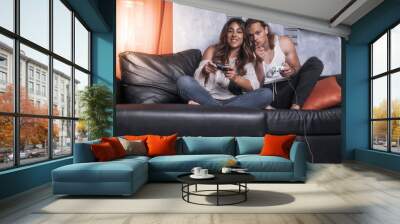 Cheerful modern couple of caucasian guys enjoy play together in living room of their studio house . Male guy enjoyng life with a girl playng videogames holding joypad and smile having fun at evening Wall mural