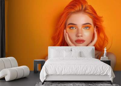 A young woman with bright orange hair and matching makeup, posing with her hands on her face against an orange background. Wall mural