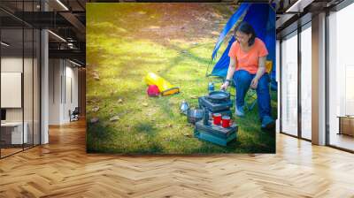 Asian woman in orange shirt and blue jean with sneakers cooking at a camping area. Outdoor kitchen equipment and yellow chair, lantern, mug and tent. Set of camping equipment with copy space. Wall mural