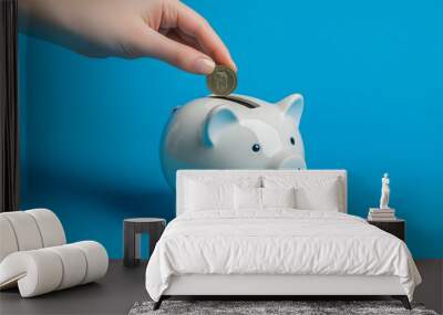 Close-up of a hand placing a coin into a piggy bank on a blue background Wall mural