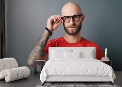 Confident young bearded man in red t-shirt fixing his black framed glasses on gray background Wall mural