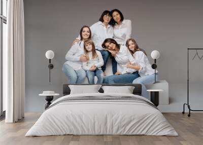 Delighted cute young girls, their moms and grandmother smiling, hugging while sitting on white cubes on gray background wearing jeans and white shirts. Lovely women group Wall mural