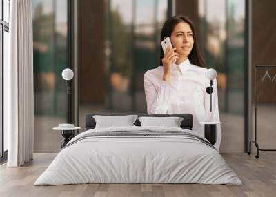 Portrait of a pretty Woman speaking on the phone wearing white business shirt. She is solving, negotiating daily tasks with her colleagues with glass walls in background. Wall mural