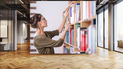 Young woman wearing glasses looking for a book on a bookshelf, i Wall mural