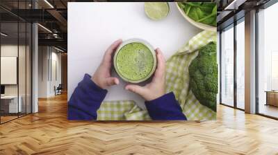 Baby is holding a freshly squeezed healthy juice of vegetables on the white wood table.  Wall mural