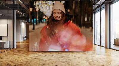 Amazing bokeh portrait of happy woman in hat and winter jacket standing posing on snow city street on blurred background of hinged bright festive illumination. Pretty female walking on urban square Wall mural